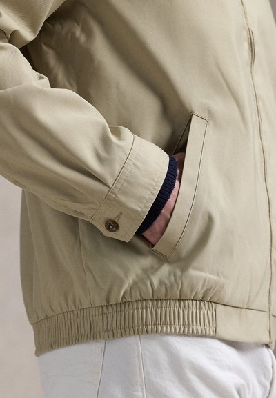 Person iført beige jakke med hånd i sidelomme, der viser elastisk talje og knaplukket manchet over mørk sweaterærme.