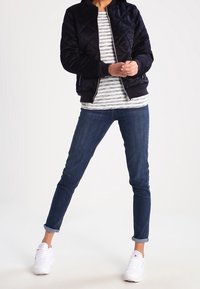 Marineblauer gesteppter Bomberjacke, gestreiftes Langarmshirt, blaue Jeans, weiße Sneakers. Die Jacke hat gerippte Bündchen und einen gerippten Kragen.