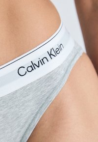 Lys grå bomullsbrief med svart "Calvin Klein" merkevare på en hvit midjebånd. Har en glatt tekstur og lavt liv.