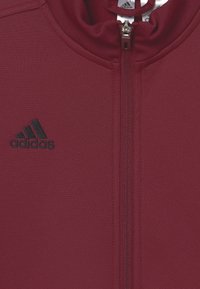 Vínově červená bunda na zip vyrobená z texturované látky, s černým logem adidas na levé straně a vysokým límcem s hladkým zipem.
