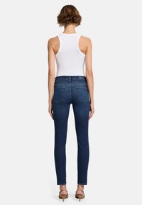 Dünne Denim-Jeans in dunkelblauer Waschung, hochgeschnitten mit Gesäßtaschen. Getragen mit einem figurbetonten weißen Tanktop und Absatzsandalen.