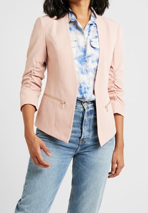 Mujer vistiendo un blazer fruncido rosa claro sobre una camisa de botones con estampado azul y blanco, combinada con vaqueros azul claro.