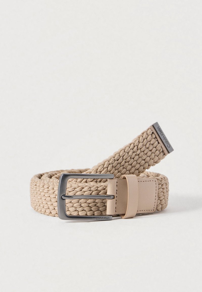 Ceinture tissée beige avec un design à sangle plate, dotée d'une boucle en métal argenté et d'un accent en cuir avec le logo de la marque.