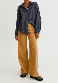 Chemise à manches longues à carreaux bleus et verts avec boutons, associée à un pantalon en velours côtelé beige taille haute à jambes larges et des bottes noires épaisses.
