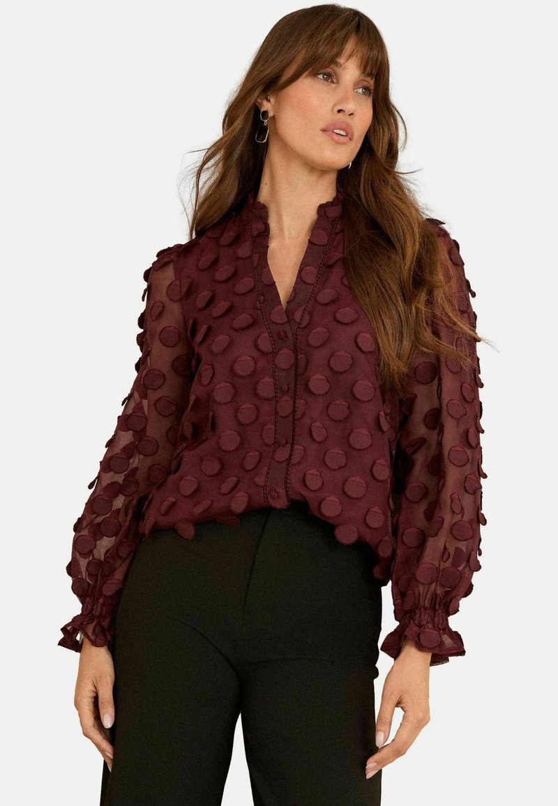 Kvinna i en genomskinlig vinröd blus med texturerade prickar och volangdetaljer vid ärmarna, ivrigt matchad med högmidjade svarta byxor.