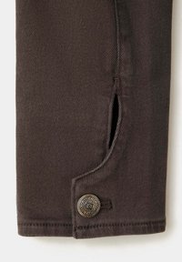 Jambe de pantalon en tissu marron avec un bouton métallique décoratif et une petite fente au-dessus de l’ourlet sur un fond clair.