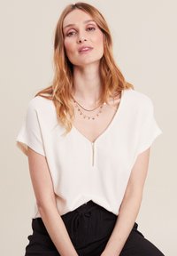Weiße kurzärmelige Bluse mit V-Ausschnitt und einem Reißverschluss vorne, aus strukturiertem Stoff. Kombiniert mit schwarzen, locker sitzenden Hosen.