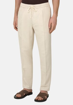 Pantalon beige pour homme avec cordon de serrage, porté avec des sandales marron, debout devant un fond clair uni.