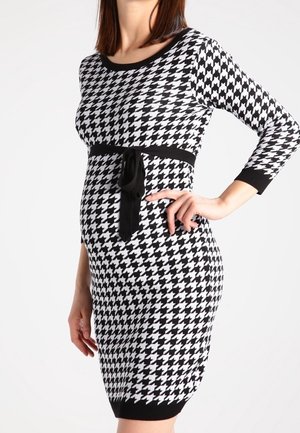 Kvinde iført kjole med sort-hvidt hundetandsmønster, trekvartlange ærmer og sort bæltetie, stående med hånden på hoften.