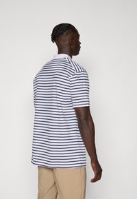 White polo shirt with navy horizontal stripes, short sleeves, and a collar. Model wearing khaki pants, viewed from the back.