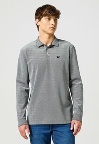 Koszula polo z długim rękawem w szarym melanżowym kolorze z kołnierzykiem, trzema guzikami i małym czarnym logo na lewej piersi. Noszona z niebieskimi dżinsami.