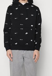 Sudadera negra con capucha que presenta logos y texto de Nike en blanco dispersos. Confeccionada con tela suave, cuenta con un bolsillo frontal y capucha con cordón ajustable.