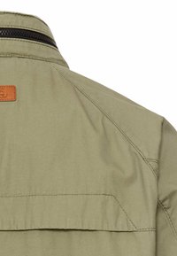 Olive green jacket featuring a zippered collar, shoulder seams, and a leather brand patch. Made of smooth fabric with a matte finish.