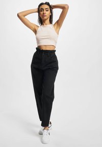Valge lühike tank top, mustad kõrge vöökohaga püksid, millel on kokkukogutud vöö, koos valgete tossudega, mudel poseerib.