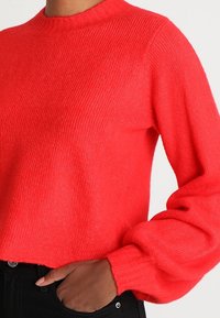Red knit sweater with a ribbed texture, round neckline, and voluminous sleeves. Paired with black pants featuring a button and pocket detail.