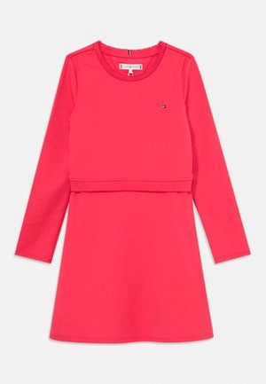 Červené šaty s dlouhým rukávem a kulatým výstřihem, s jemným detailem švu v pase a malým logem na levé horní straně hrudi.