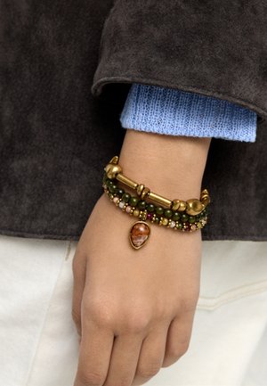 Nadgarstek z warstwowymi bransoletkami z zielonymi koralikami, złotymi akcentami i brązowym wiszącym w kształcie kropli, na niebieskim dzianinowym rękawie i ciemnej kurtce.
