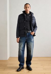 Jachetă de exterior bleumarin cu multiple buzunare, purtată peste un top negru cu fermoar, combinată cu blugi deschiși, decolorati, și cizme negre până la gleznă.