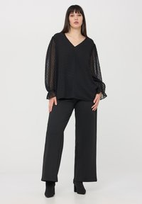 Blusa nera con motivo a pois testurizzati e scollatura a V, abbinata a pantaloni neri a gamba larga. Maniche a sbuffo trasparenti con polsini elasticizzati.