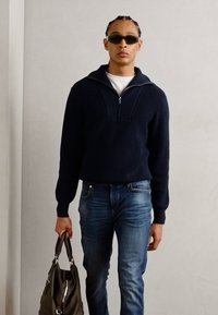 Marineblå strikket sweater med lynlås krave, blå jeans og en olivengrøn taske. Glat tekstur og moderne silhuet.