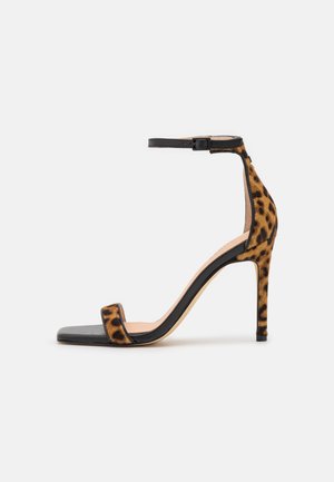 Sandal de tacón con tira al tobillo en estampado de leopardo y base de cuero negro. Presenta un tacón elegante y delgado, así como un diseño minimalista.