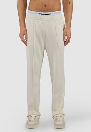 Hombre vistiendo pantalones de pierna recta color crema, zapatillas blancas y ropa interior con cintura elástica blanca con el logo "PEGADOR", de pie contra un fondo liso.