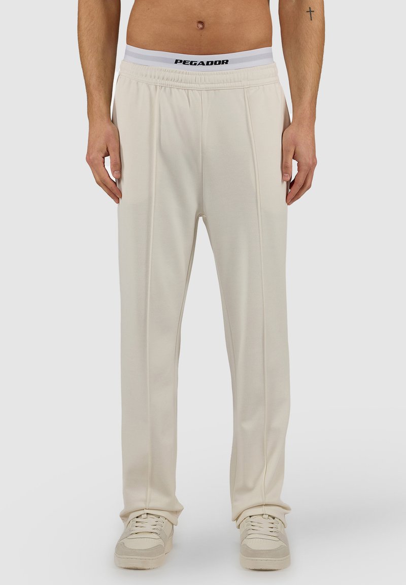 Mies, joka pukeutunut luonnonvalkoisiin suoria lahkeita oleviin housuihin, valkoisiin tennareihin ja valkoisiin, joustavaan vyötärönauhaan, jossa on "PEGADOR"-logo, oleviin alushousuihin, seisoo yksivärisen taustan edessä.