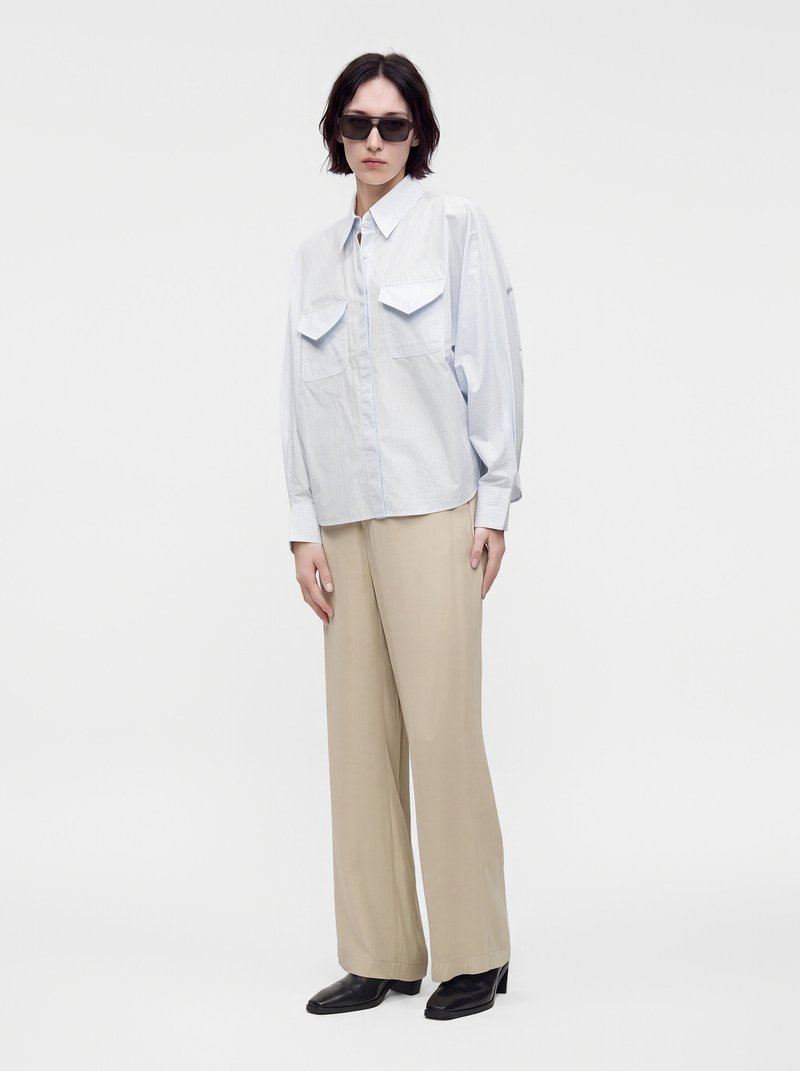 Femme portant des lunettes de soleil noires, une chemise bleu clair oversize avec des poches poitrine, un pantalon beige à jambes larges et des chaussures à talons noires, debout sur fond blanc.