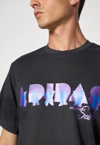 Sort bomulds T-shirt med dristig lilla og blå grafisk tekst, der staver "ADIDAS" med silhuetter og geometriske former. "JEREMY SCOTT" logo inkluderet.
