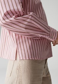 Jasnoróżowa koszula w paski z brązowymi liniami, lekka tkanina, mankiety na rękawach, rozcięcia po bokach i guziki przy kołnierzu. W połączeniu z beżowymi spodniami.
