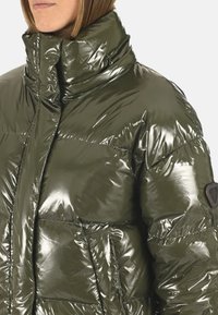 Doudoune brillant olive verte avec un col haut, des coutures visibles, un matelassage horizontal et un détail de patch sur la manche gauche.