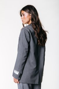 Ženska z dolgimi valovitimi lasmi nosi sivo jakno s pinstripes vzorcem, na kateri je oznaka na rokavu, obrnjena levo v bočni profil proti beli ozadju.
