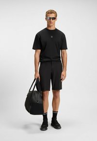 Czarna koszulka sportowa z logo, czarne szorty, czarne skarpetki i okulary przeciwsłoneczne. Trzyma czarną torbę sportową z paskiem. Matowe tekstury w całej kompozycji.