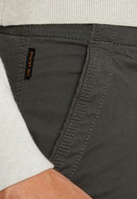 Pantalons en tissu gris avec une poche latérale ornée d'une petite étiquette noire portant l'inscription "Pine Legend." Détails de couture visibles et texture douce.