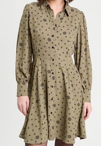 Femme portant une robe boutonnée olive avec des motifs floraux noirs et de longues manches bouffantes, se tenant devant un fond uni.