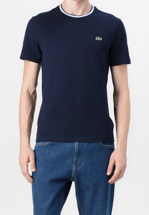 Man draagt een marineblauwe T-shirt met korte mouwen en een klein groen krokodillenlogo op de borst, gecombineerd met een blauwe spijkerbroek.