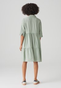 Knielanges, hellgrünes Kleid aus strukturiertem Stoff, mit kurzen Ärmeln und gestuftem Design. Kombiniert mit weißen Turnschuhen mit marineblauen Streifen.