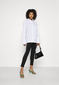 Camicia bianca oversize con sottili righe blu, leggings neri con righe laterali, sandali verdi con tacco e una piccola borsa nera.