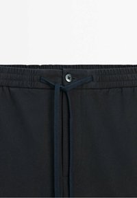 Taille de pantalon noire avec bande élastique, bouton noir et cordon noué centré sur fond uni.