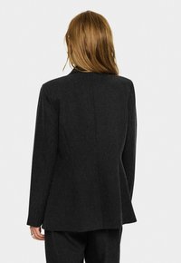Donna con lunghi capelli castano chiaro che indossa un blazer nero strutturato e pantaloni abbinati, vista da dietro su uno sfondo semplice.