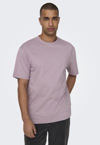 Camiseta de manga corta de color púrpura claro con cuello redondo, hecha de algodón, que presenta un ajuste holgado y una textura lisa. Lleva puesta un pantalón gris oscuro.