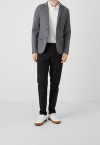 Blazer grigio texturizzato su una polo bianca, abbinato a pantaloni neri e sneaker bianche con suole marrone chiaro.