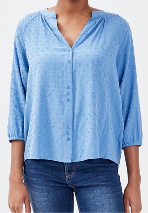 Blouse bleu clair à boutons avec des points texturés, manches 3/4, associée à un jean bleu foncé.