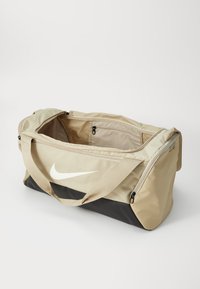 Beige duffeltas met een zwarte bodem, groot Nike-logo, dubbele ritsen en twee bovenhandvatten voor gemakkelijk dragen. Interieur met ritsvak.