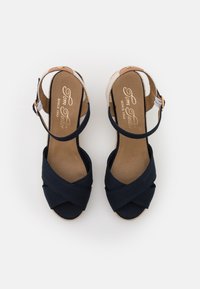 Sandales espadrilles bleu marine avec brides croisées, bride de cheville beige et talon compensé en espadrille texturé. Semelle intérieure douce avec marquage visible.