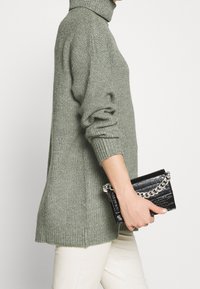 Suéter verde de punto acanalado con un cuello alto, combinado con un clutch negro de piel sintética que cuenta con una cadena plateada.