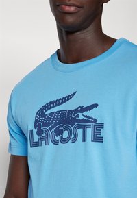 Mand iført en lyseblå T-shirt med et stort mørkeblåt krokodillelogo og stiliseret "Lacoste" tekst over brystet.