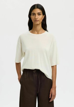 Femme aux longs cheveux noirs portant un haut blanc ample à manches courtes et un pantalon marron foncé à cordon, debout devant un fond clair uni.