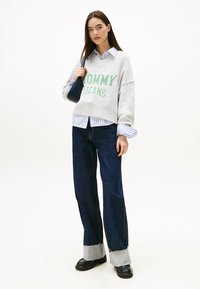Grauer Strickpullover mit "MOMMY JEANS" in Grün, über einem blau gestreiften Hemd getragen. Dunkelblaue weit geschnittene Jeans mit fransigem Saum und schwarzen Schuhen.