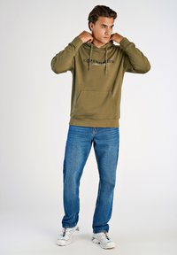 Olivgrön hoodie med en framficka och snodd, med "KÖPENHAMN" tryckt i marinblått. I kombination med blå denimjeans och vita sneakers.